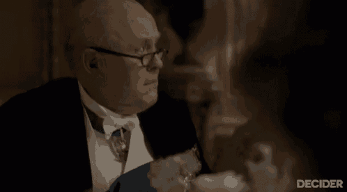 a man in a tuxedo and glasses is sitting at a table talking to a woman .