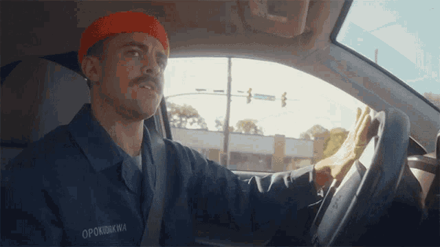 a man wearing an orange beanie and a blue shirt with the name opokukwa on it is driving a car
