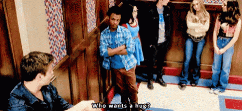 a group of people are standing in a hallway and one of them says who wants a hug .