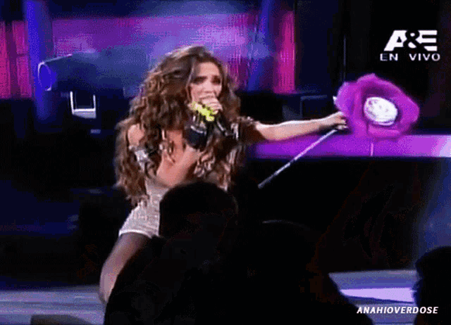a woman singing into a microphone while holding a purple stuffed animal with the letters ae on the bottom right