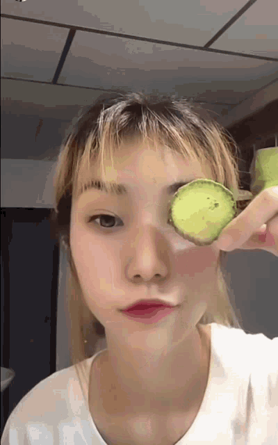 a woman holding a slice of cucumber over her eye