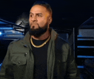 a man with a beard and a gold chain is wearing a green jacket and black shirt .