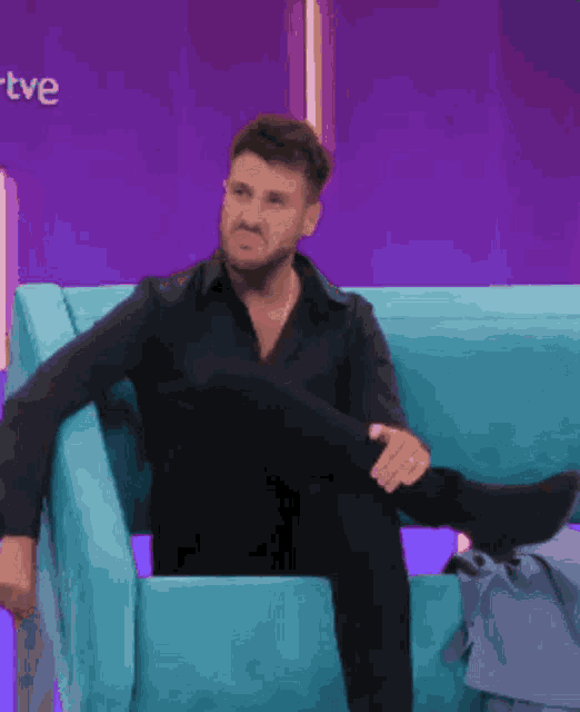 a man in a black shirt sits on a blue couch with his leg up