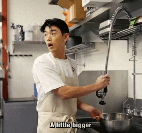 a man in an apron is standing in a kitchen with the words a little bigger above him