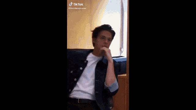 a man in a denim jacket is sitting in a chair with his hand up .