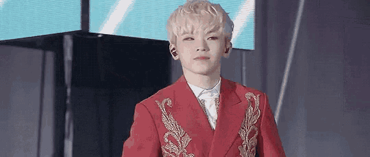 a young man wearing a red jacket and a white shirt is standing in front of a large screen .