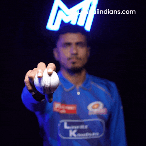 a man in a blue shirt is holding a cricket ball in front of an indians.com sign