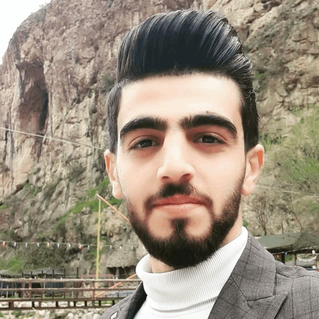 a man with a beard wearing a suit and a white turtleneck poses for a picture