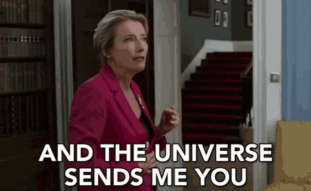 a woman in a pink jacket is standing in a living room and the universe sends me you