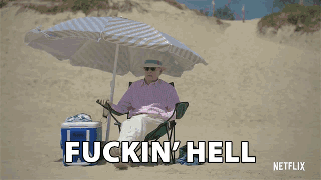 a man sits under an umbrella on a beach and says fuckin hell