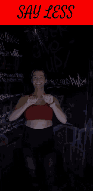 a woman in a red top and black shorts stands in front of a sign that says " say less "