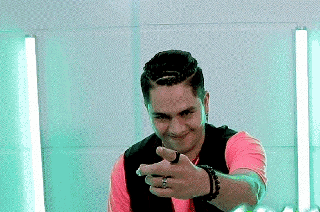 a man wearing a pink shirt and black vest points at the camera