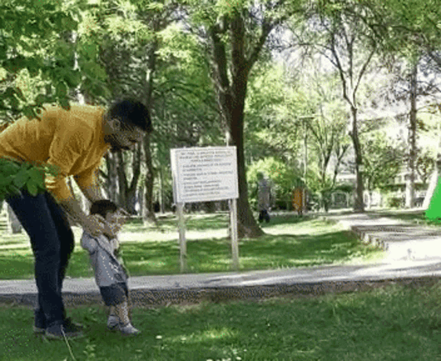 a man is holding a baby in a park with a sign in the background .