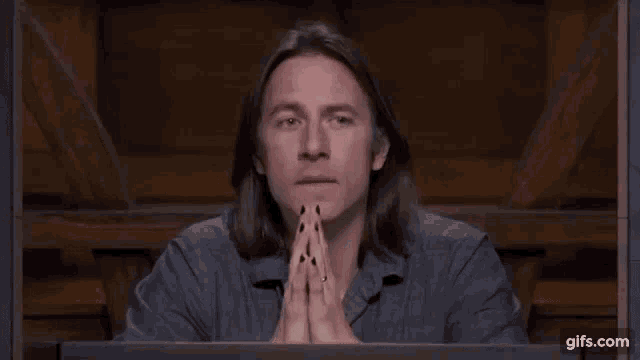 a man with long hair and black nails is sitting at a table with his hands folded .