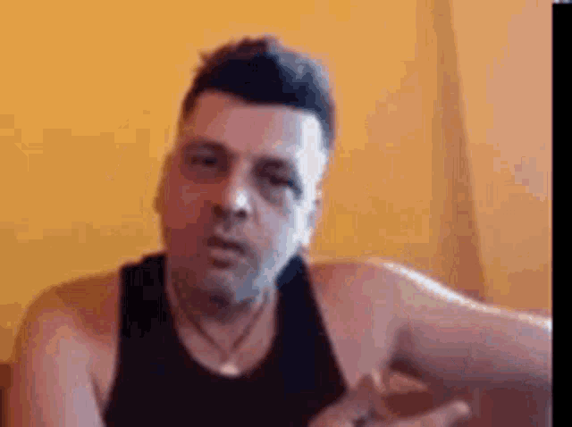 a man in a black tank top is sitting in front of a yellow wall and making a funny face .