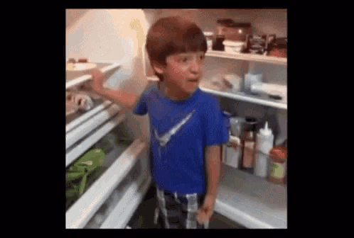 a young boy in a blue nike shirt is standing in a refrigerator .