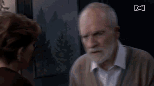 a man with a beard is talking to a woman in front of a tv screen with the letters bc on it
