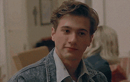 a young man wearing a denim jacket and a white shirt is sitting at a table looking at the camera .