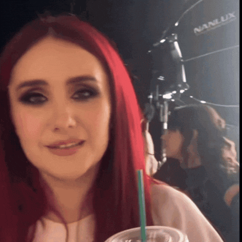 a woman with red hair is holding a drink in front of a nan lux light