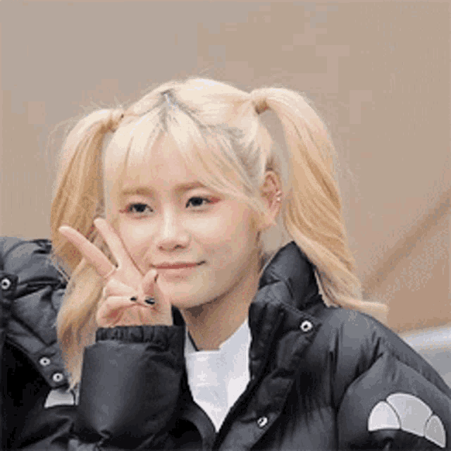 a girl with blonde hair and pigtails is wearing a black jacket and giving a peace sign