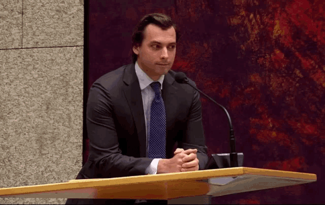 a man in a suit and tie is sitting at a podium with his hands folded