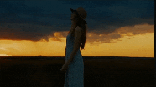 a woman in a white dress and hat is standing in a field at sunset