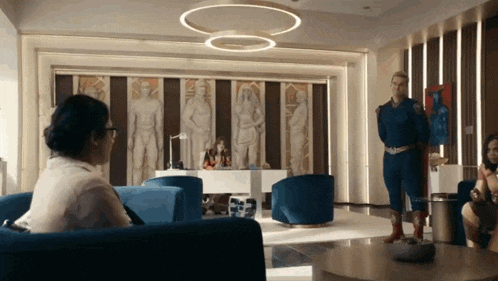 a man in a superhero costume is standing in a living room with a woman sitting in a chair .