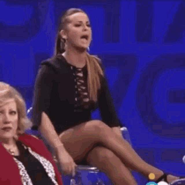 a woman in a black dress is sitting on a chair with her legs crossed and waving .