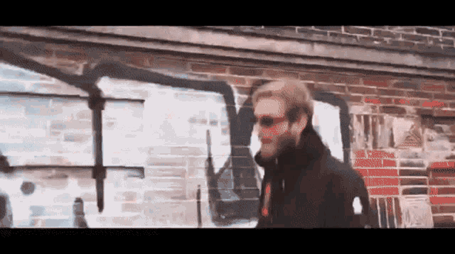 a man wearing sunglasses is standing in front of a brick wall with graffiti on it including the letter t