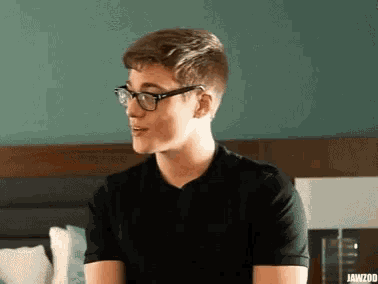 a young man wearing glasses and a black shirt is sitting on a bed with jawzoo written on the bottom right