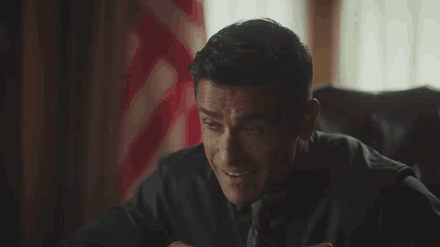a man in a suit and tie is smiling while sitting in front of a flag