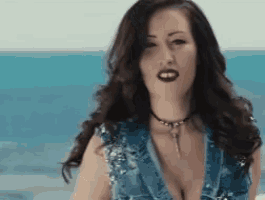 a woman wearing a choker and a denim vest is standing on a beach looking at the camera .