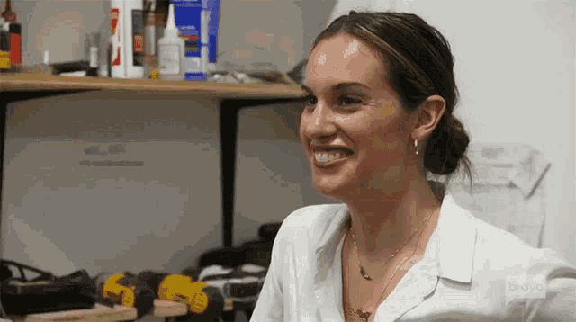 a woman in a white shirt is smiling while sitting in front of a shelf with tools .