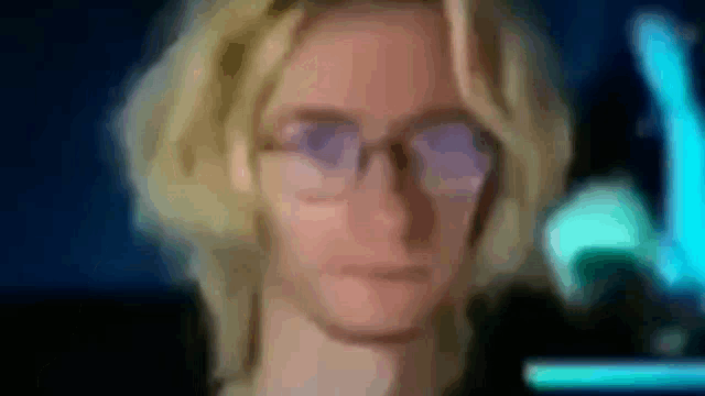 a man with blonde hair and glasses is looking at the camera in a blurry photo .