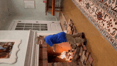 a boy in a blue shirt is standing in front of a fireplace with a picture of a family on the wall behind him