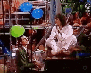 a man is sitting on a stage next to a woman singing into a microphone while holding balloons .