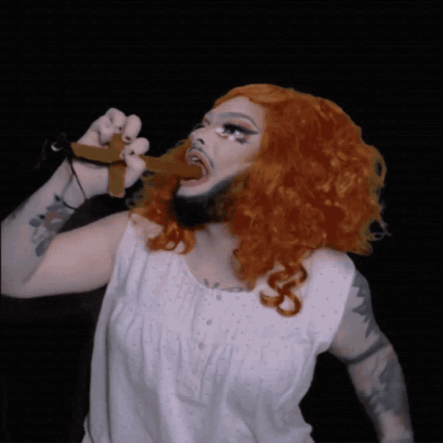 a drag queen with red hair and a beard holds a cross in her mouth