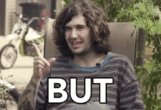 a man with curly hair is sitting in a chair with the word but written on his shirt .