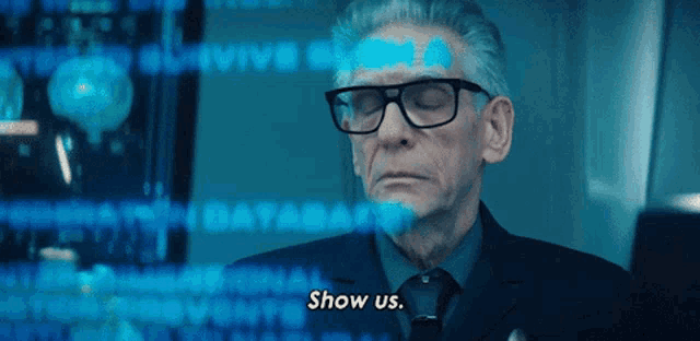 a man wearing glasses says show us in front of a screen