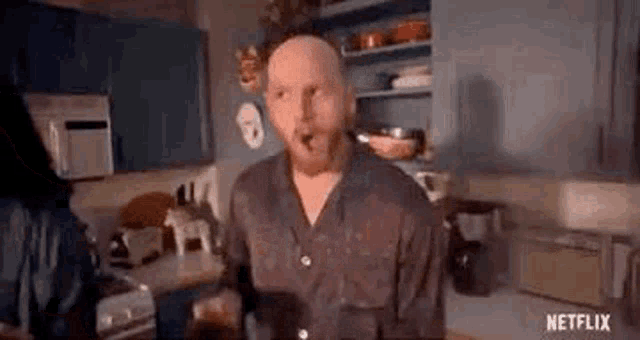 a bald man with a beard is standing in a kitchen eating a sandwich .