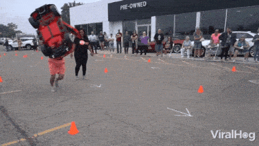 a man is carrying a toy car on his head in front of a pre-owned car dealership