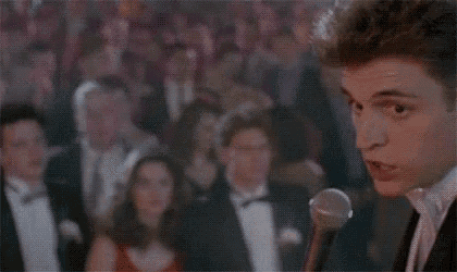 a man in a tuxedo is speaking into a microphone in front of a crowd of people .