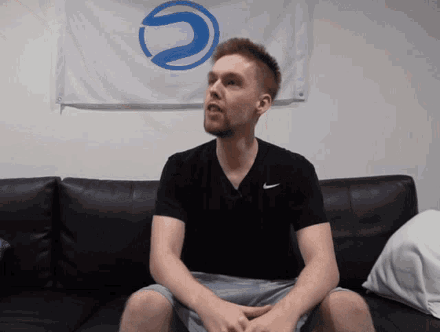 a man wearing a black nike shirt sits on a couch in front of a blue and white flag
