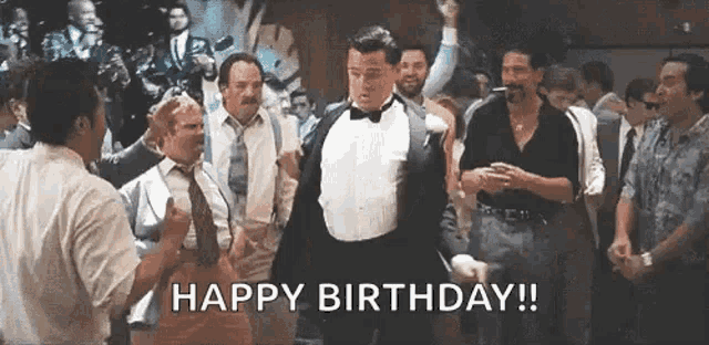 a man in a tuxedo is dancing in front of a crowd of men and says `` happy birthday '' .