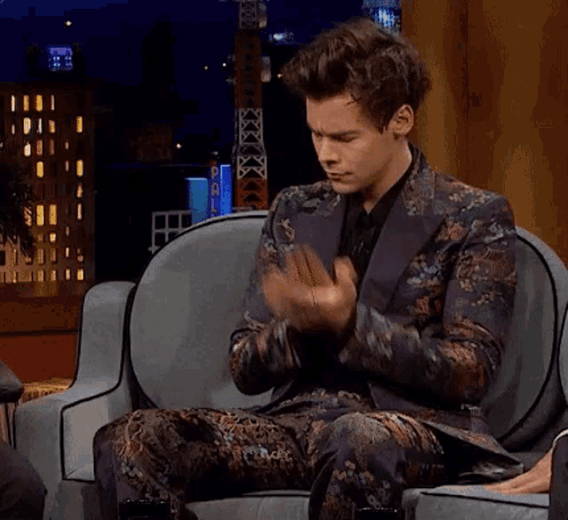 a man in a floral suit is sitting in a chair with his hands folded in prayer .