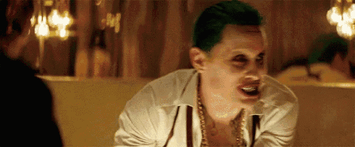 the joker is wearing a white shirt and a gold necklace while talking to a woman .