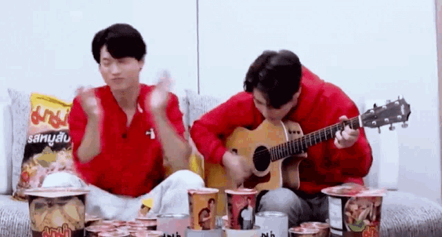 two men are playing guitars while sitting on a couch surrounded by cups of food .