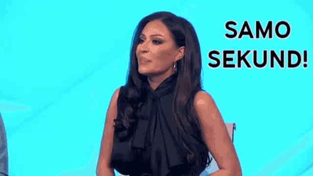 a woman in a black dress is sitting in front of a blue background and pointing her finger at something .