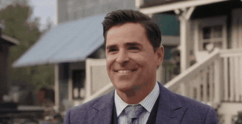 a man in a suit and tie smiles in front of a house .