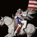 a man in a cowboy hat is riding a white horse and holding an american flag .